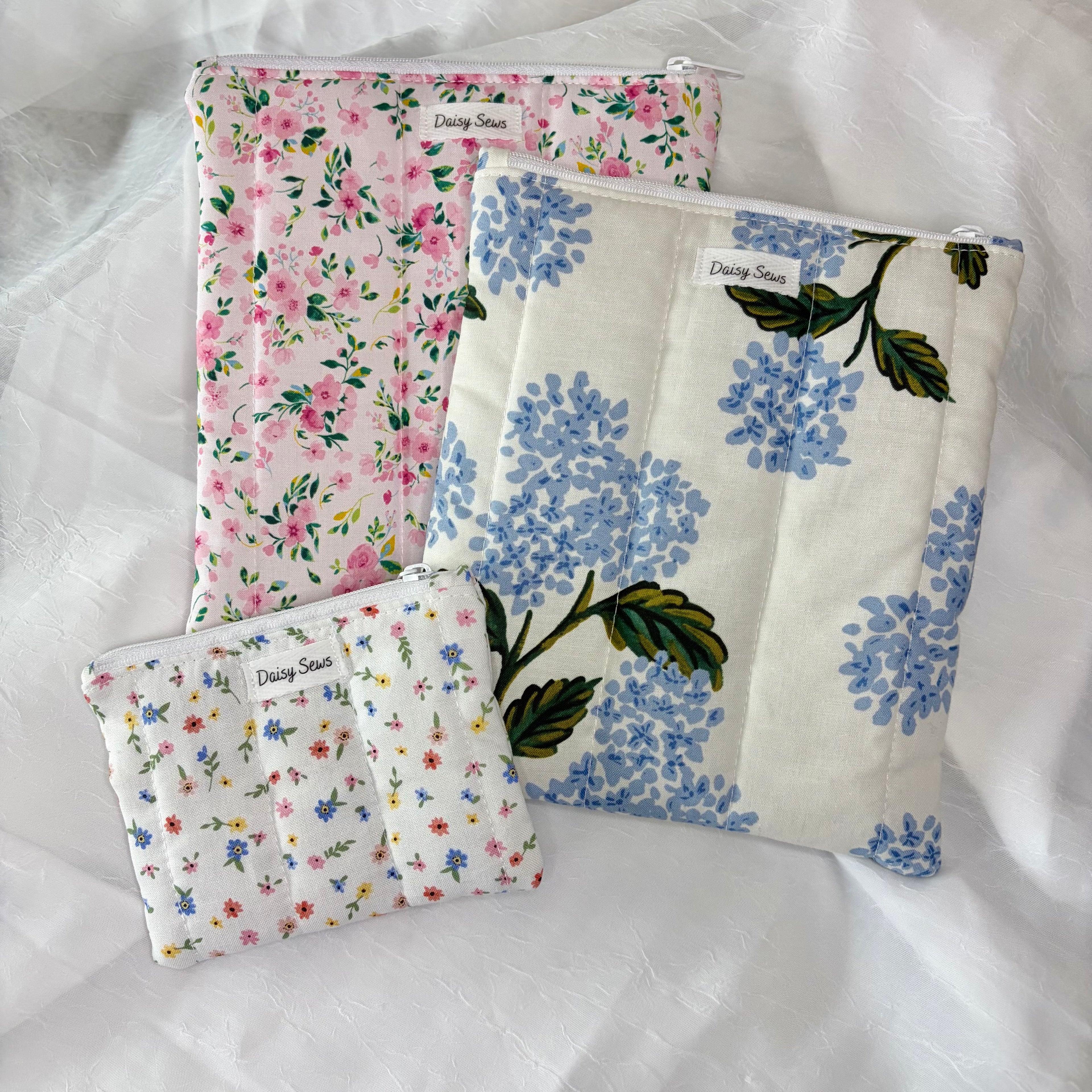 three flat pouches on white background. small pouch with little colorful flowers. Large plush pouch with blue hydrangea and green leaves and another with pink flowers