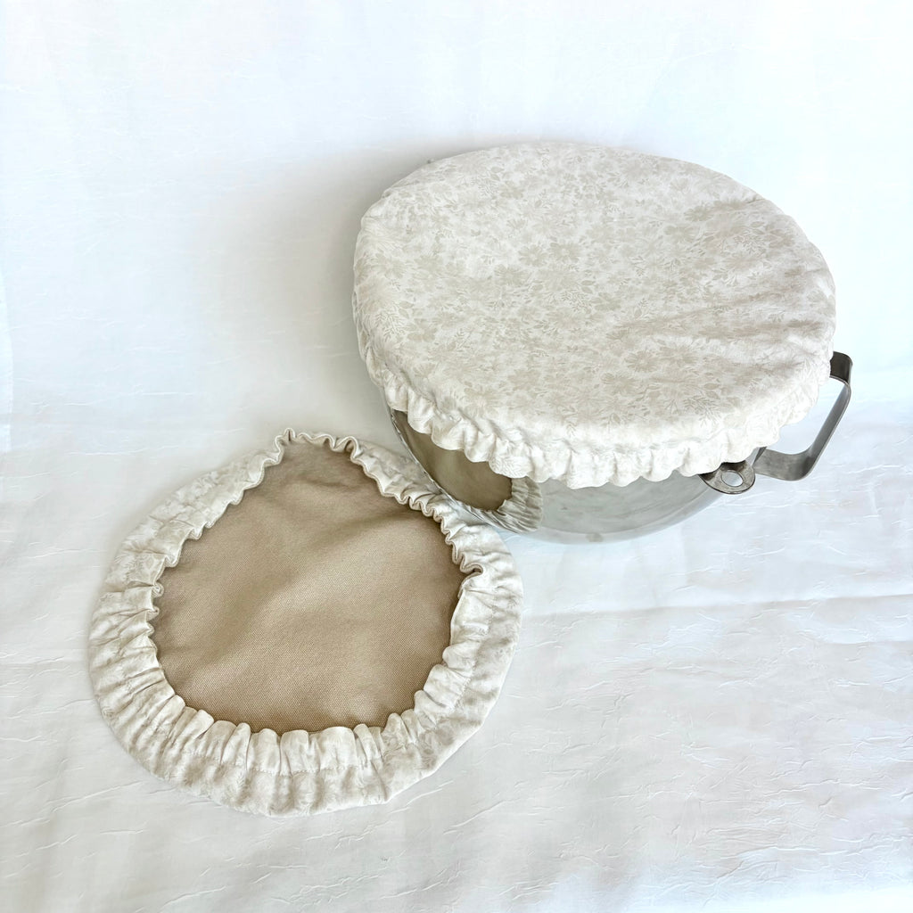 A fabric bowl cover set in cream and beige, featuring a textured cream velvet-like top cover with a ruffled edge and a matching beige inner lining, shown on a white fabric background.