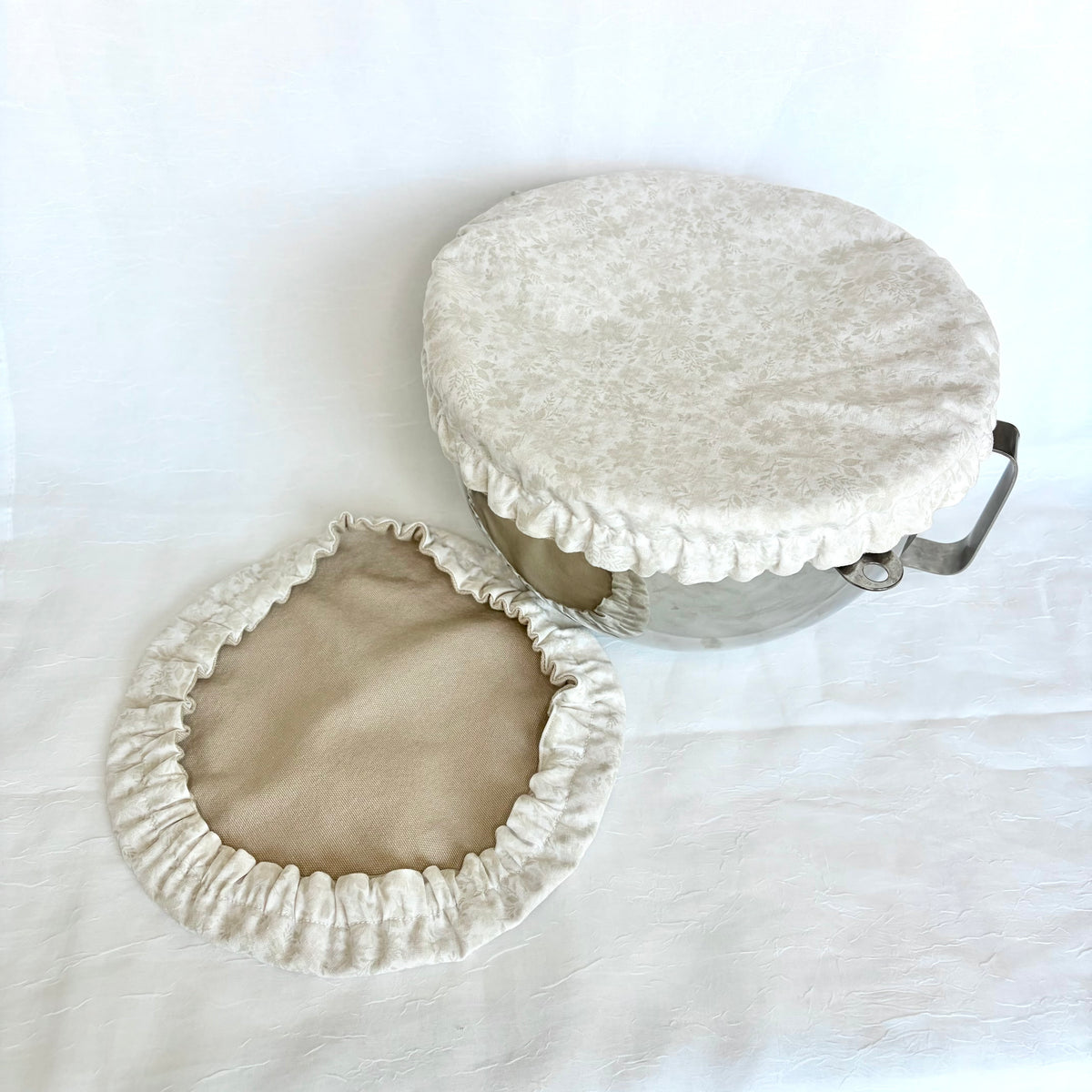 A fabric bowl cover set in cream and beige, featuring a textured cream velvet-like top cover with a ruffled edge and a matching beige inner lining, shown on a white fabric background.