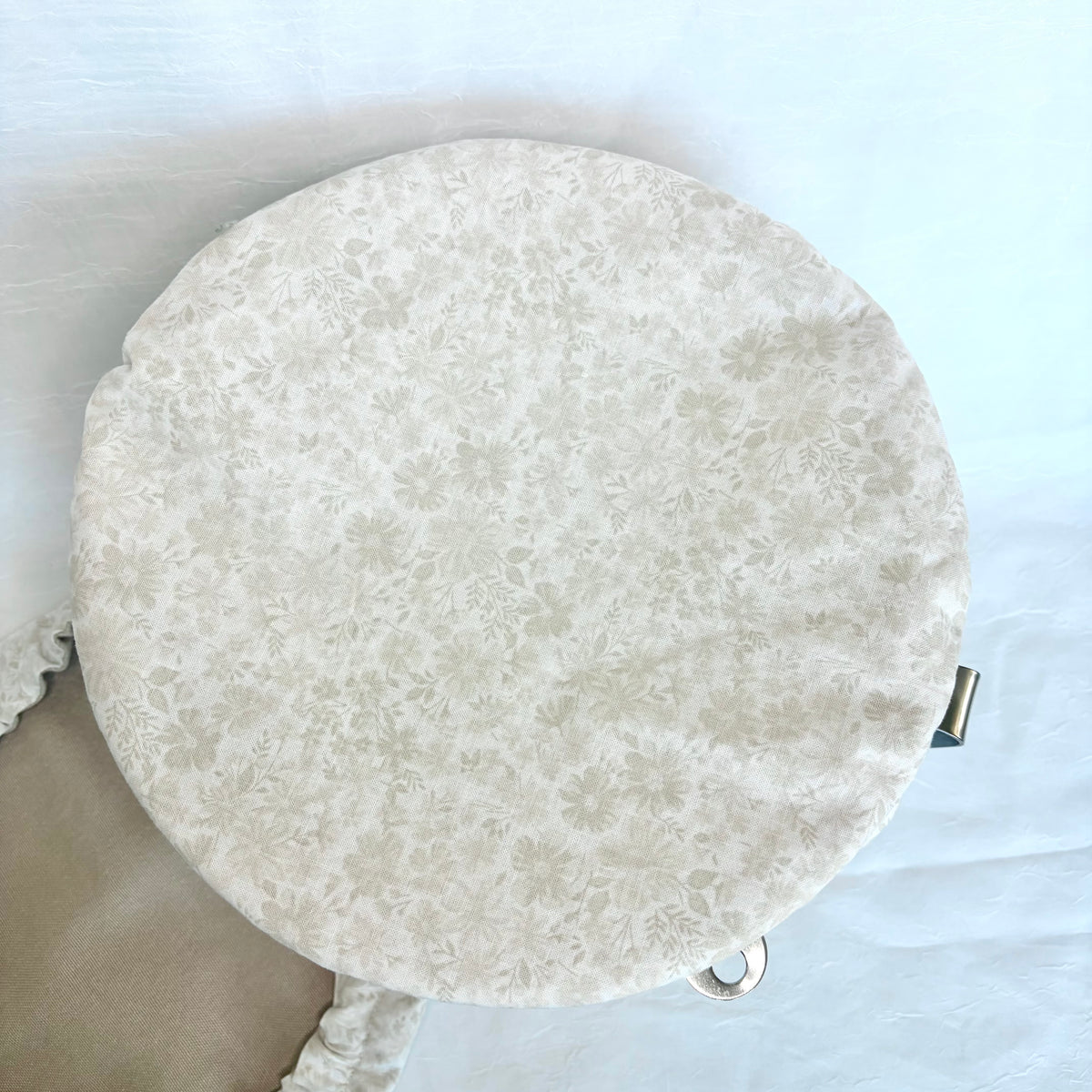 A round, off-white fabric seat cushion with a delicate beige floral pattern, shown on a light blue background with a small metal ring attachment visible on the right side.