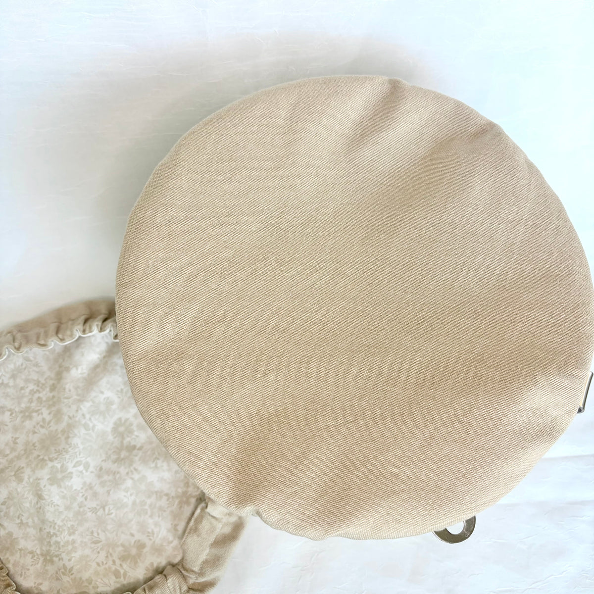 A round bowl cover in a light beige linen-like fabric, photographed from above on a white background. Part of a matching patterned cushion is visible at the lower left.