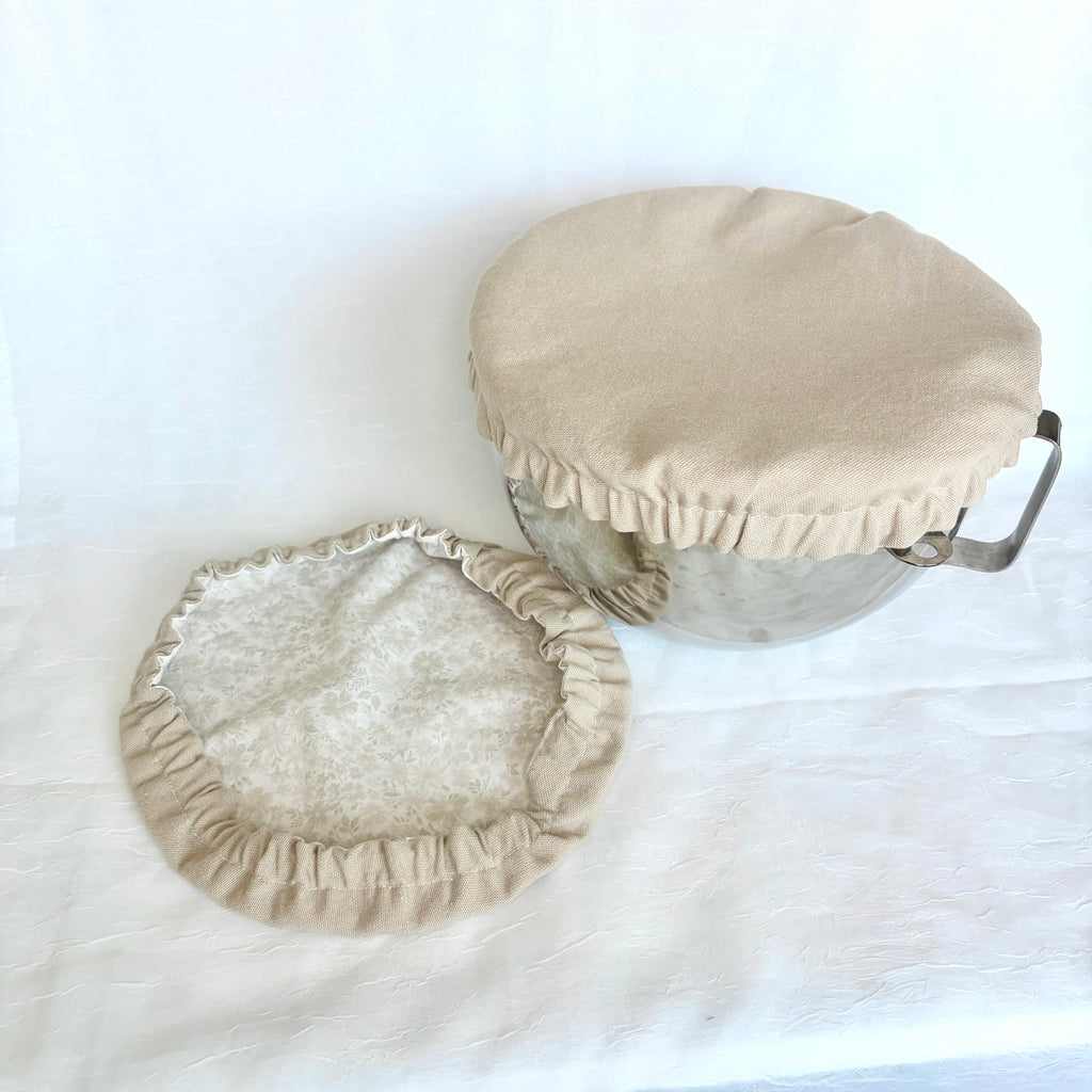 A beige fabric pot cover with a ruffled edge shown on a metal bowl, with the matching cover laid flat beside it on a white fabric background.