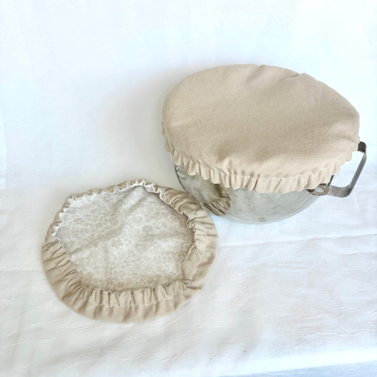 A beige fabric pot cover with a ruffled edge shown on a metal bowl, with the matching cover laid flat beside it on a white fabric background.