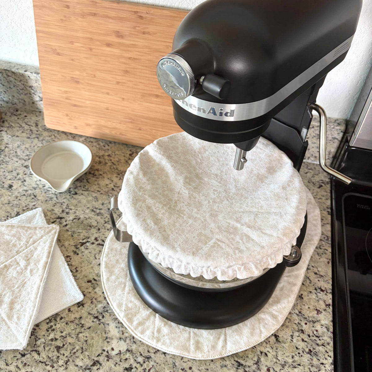 A black KitchenAid stand mixer with a white textured fabric dust cover on the bowl, set on a granite kitchen countertop next to a wooden cutting board and a small white dish.