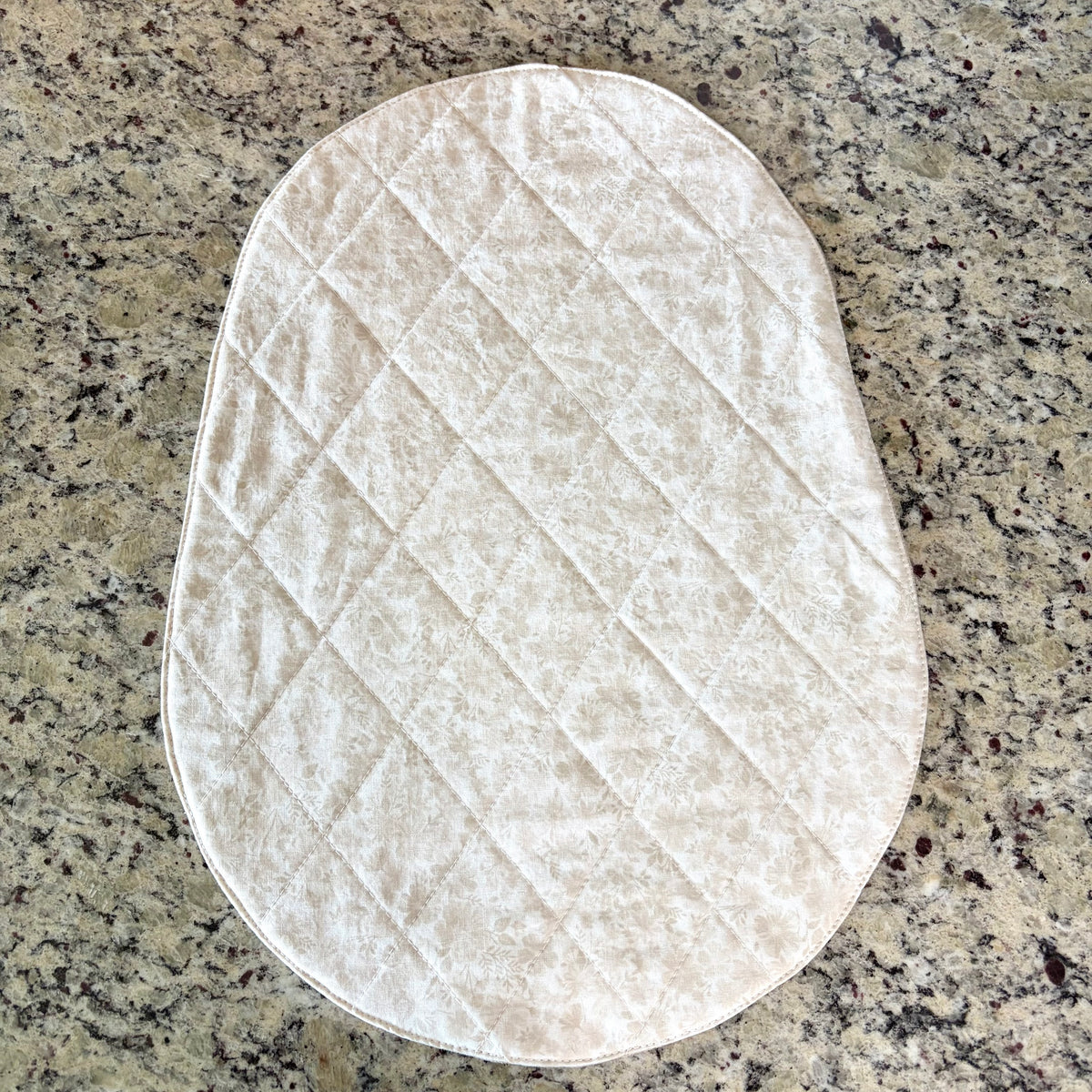 An oval quilted mat in a cream/off-white floral textured fabric, laid flat on a speckled granite countertop. Bowl Lift Size