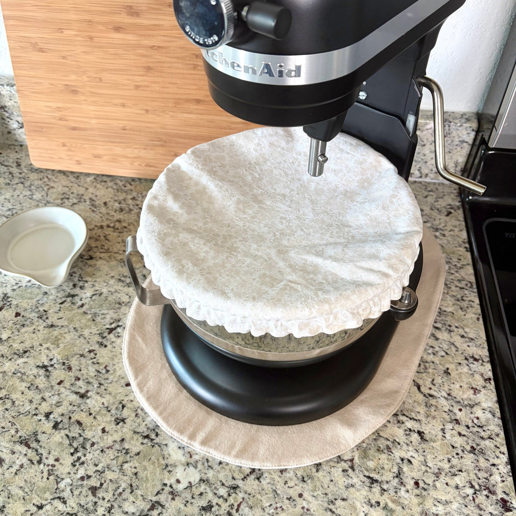 A black KitchenAid stand mixer with a beige fabric dust cover on the bowl, set on a tan mat on a granite kitchen countertop.