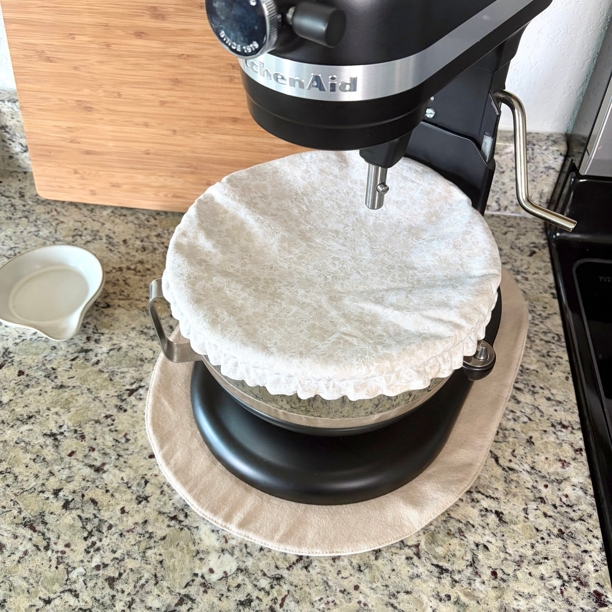 A black KitchenAid stand mixer with a beige fabric dust cover on the bowl, set on a tan mat on a granite kitchen countertop.