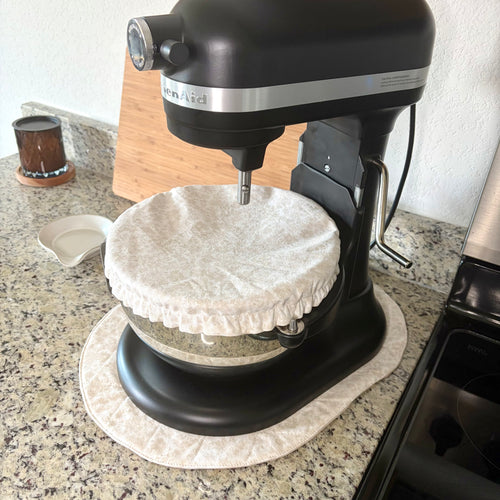 A black KitchenAid stand mixer with a fabric-covered bowl sits on a granite kitchen countertop. A matching round fabric pad is placed underneath the mixer base, with a small dish and a wooden cutting board nearby on the counter.