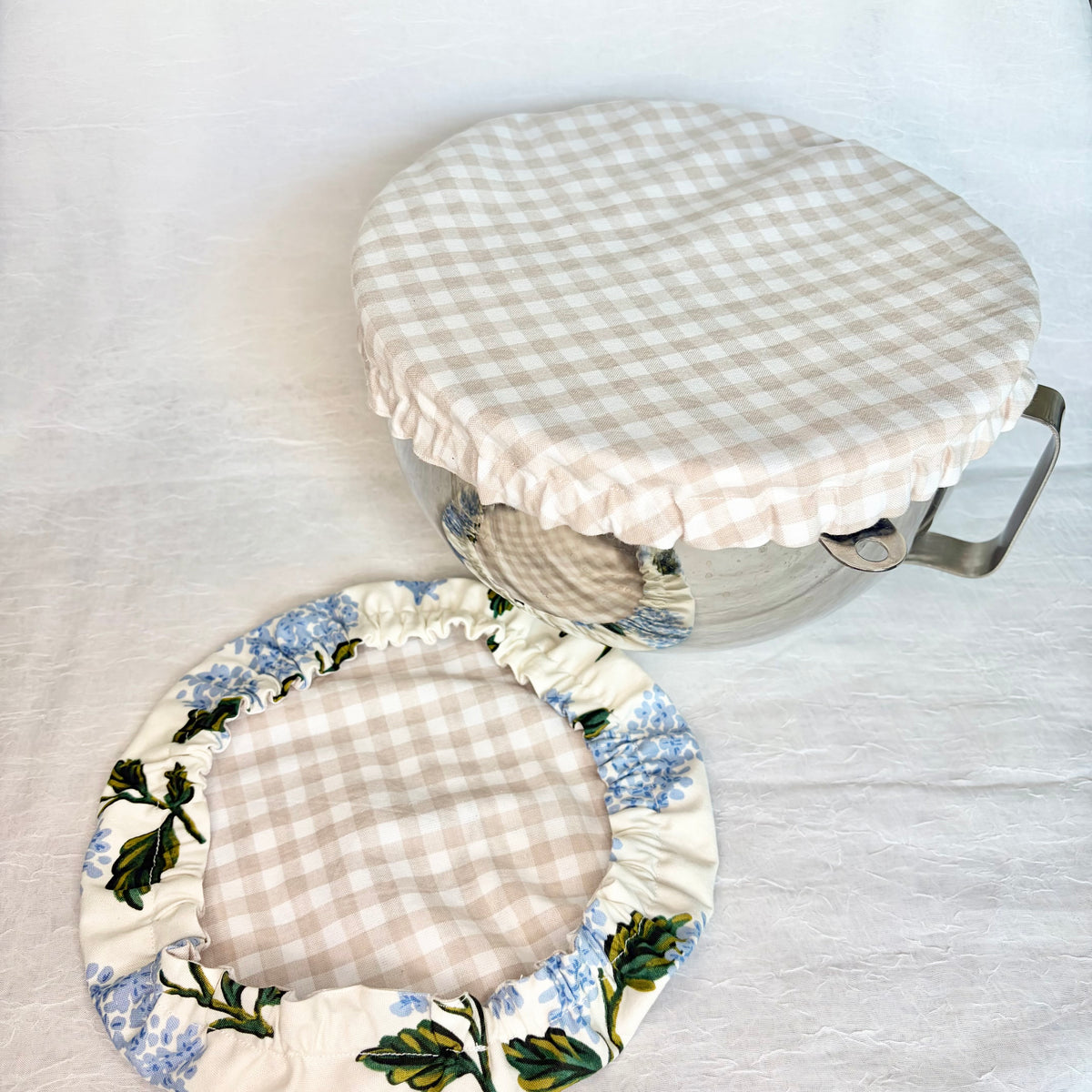 Beige Gingham fabric bowl cover displayed on a metal bowl with a handle and a matching cover with beige gingham interior and Blue hydrangea with green leaves exterior on a light gray background