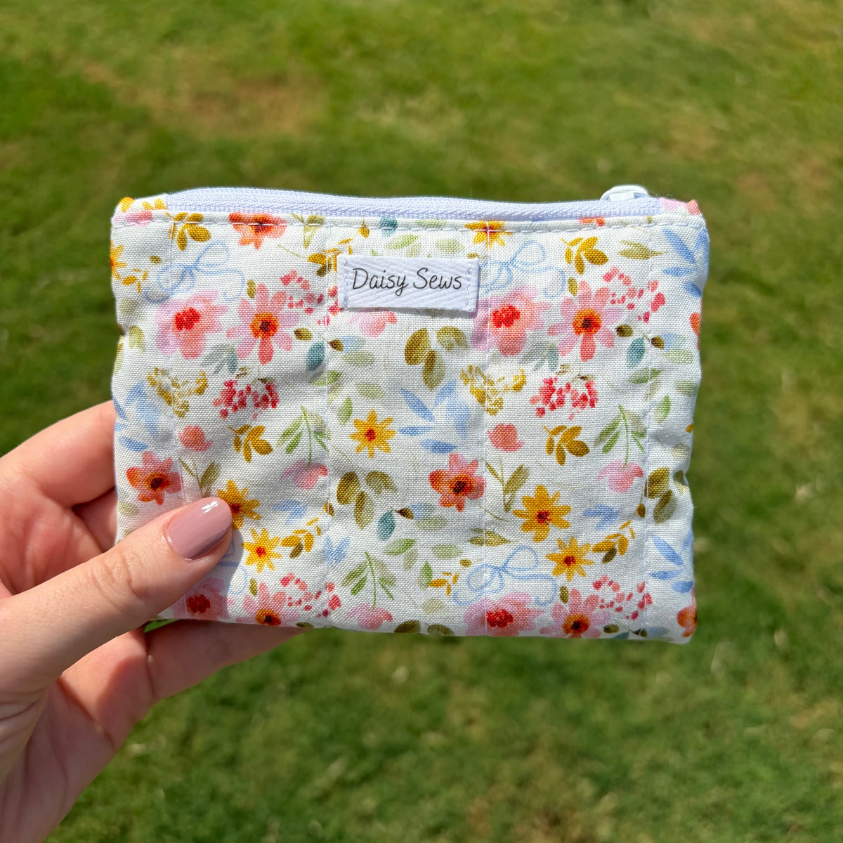A hand holds a small zippered pouch with a light blue background and a colorful floral print. The pouch has a white fabric label with the brand name 'Daisy Sews' printed on it, and the background is a blurred green grassy field.