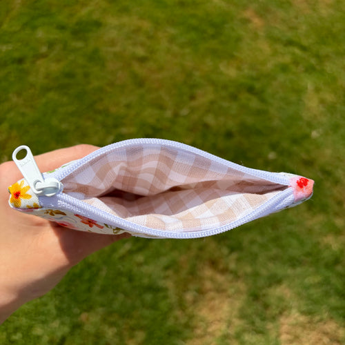 A hand holding an open zippered pouch with a white and beige gingham patterned fabric exterior and floral lining, set against a blurred green grass background.
