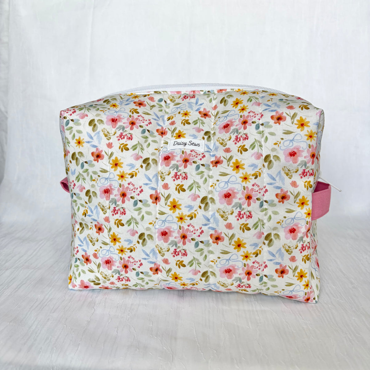 A rectangular zippered pouch with a colorful floral print on a cream background, featuring pink handles on the sides and a small embroidered 'Daisy Sews' label on the front.