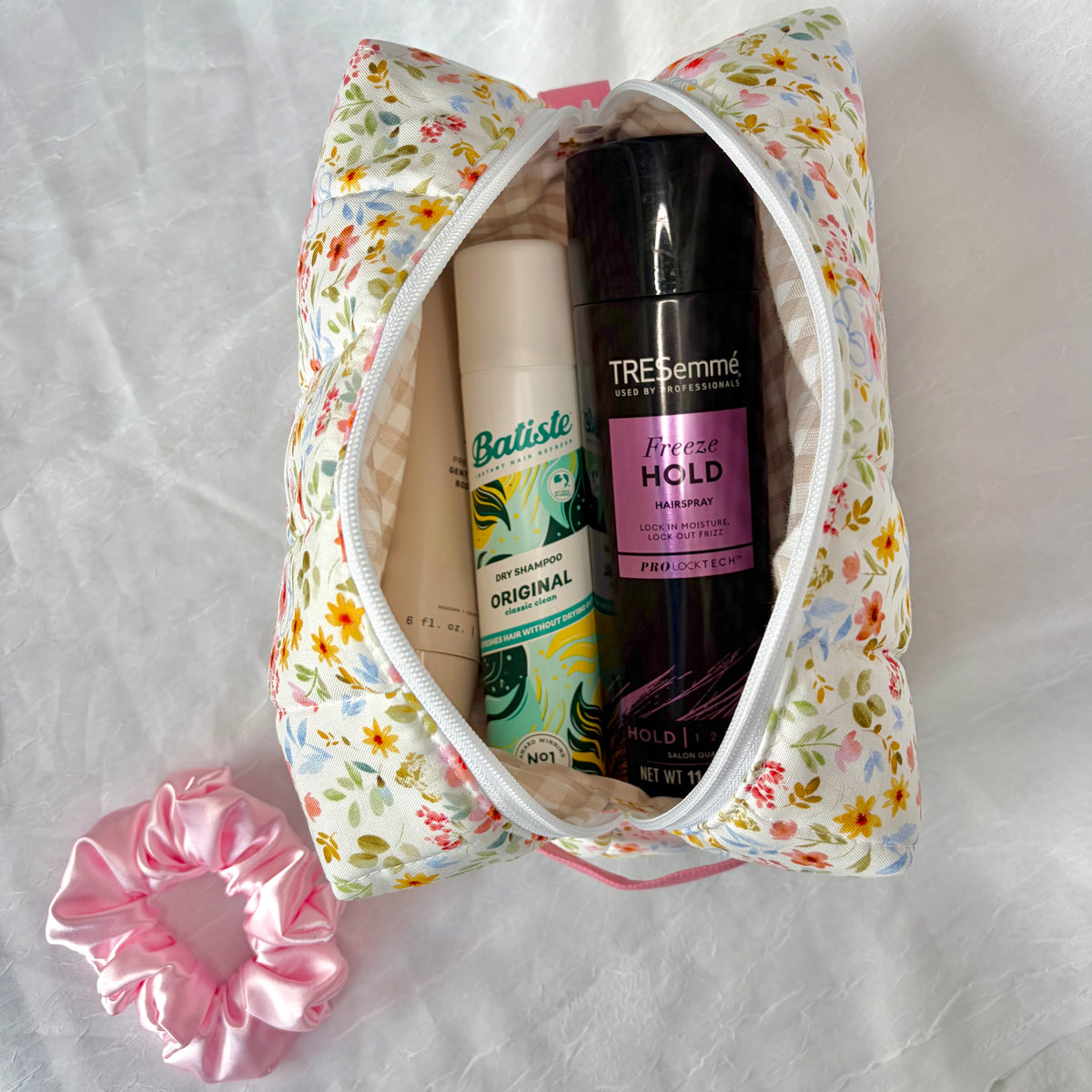 A floral zippered cosmetic pouch filled with hair care products, including a Tresemme Freeze Hold hairspray and a Batiste Original dry shampoo, on a white fabric background with a pink scrunchie.
