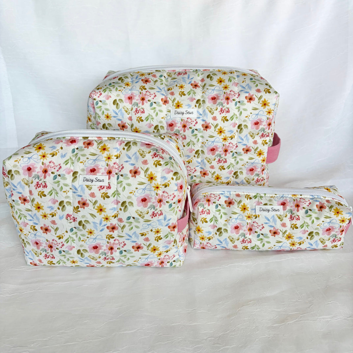 Three floral-print zippered pouches in different sizes arranged on a white fabric backdrop. The pouches have a delicate multicolor floral pattern with pink, yellow, and green flowers on a light background, with a small white label reading 'Daisy Sews' on each.