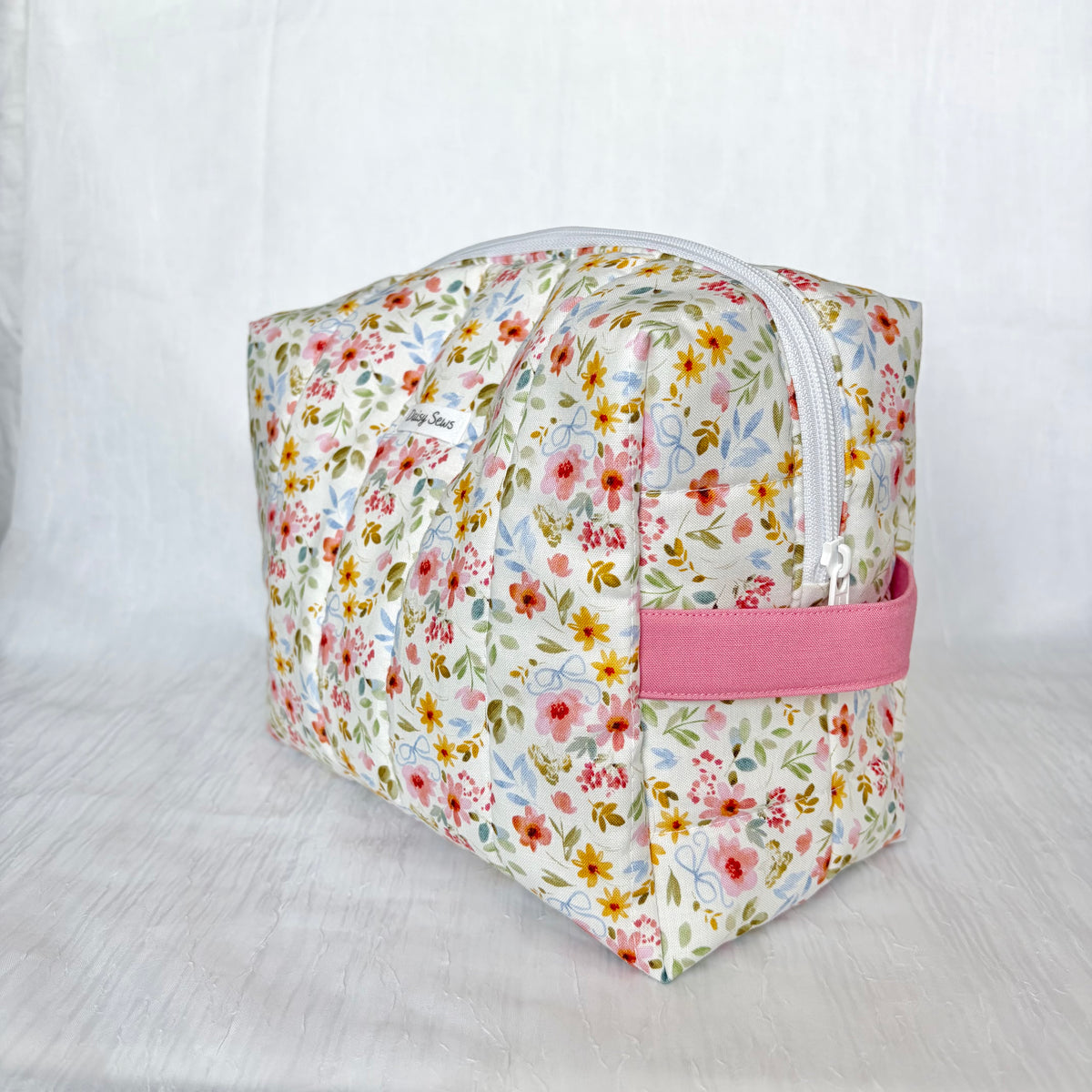 A rectangular zippered toiletry bag with a white background and a colorful floral print, featuring a pink fabric handle on the side.