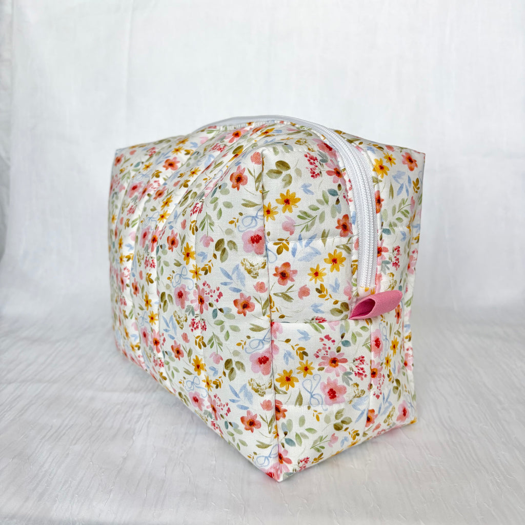 A rectangular zippered pouch with a white background and a dense multicolor floral print featuring pink, red, yellow, and green flowers. It has a pink pull tab and is photographed on a white fabric backdrop.