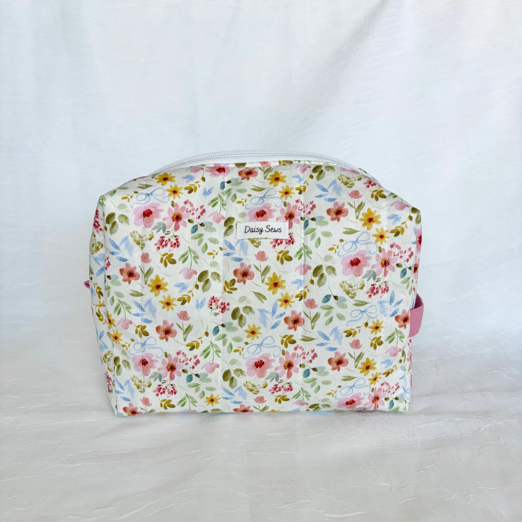 A small rectangular cosmetic pouch with a white background and a dense multicolor floral print featuring pink, red, yellow, and green flowers with blue leaves. A small white label on the front reads 'Daisy Sews'.