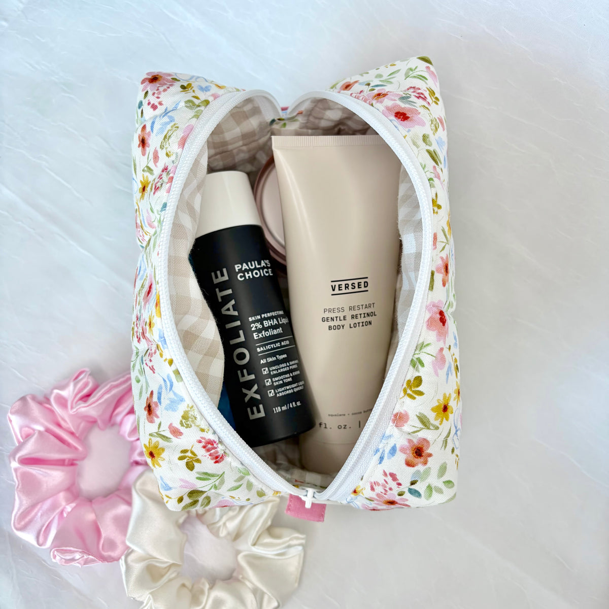 A floral zippered cosmetic pouch filled with skincare products, including a black PAULA CHOICE EXFOLIATE bottle and a beige VERSED body lotion tube, styled on a white textured background with pink and cream satin scrunchies.
