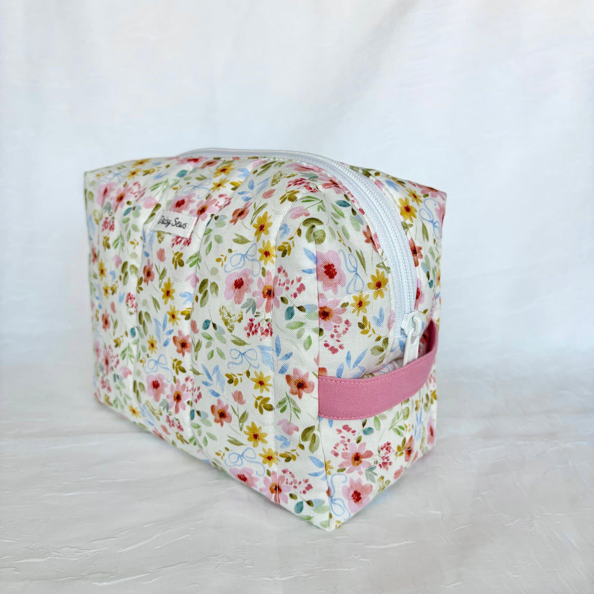 A rectangular zippered cosmetic pouch with a white background and a colorful floral print, featuring a pink fabric loop handle on the side.