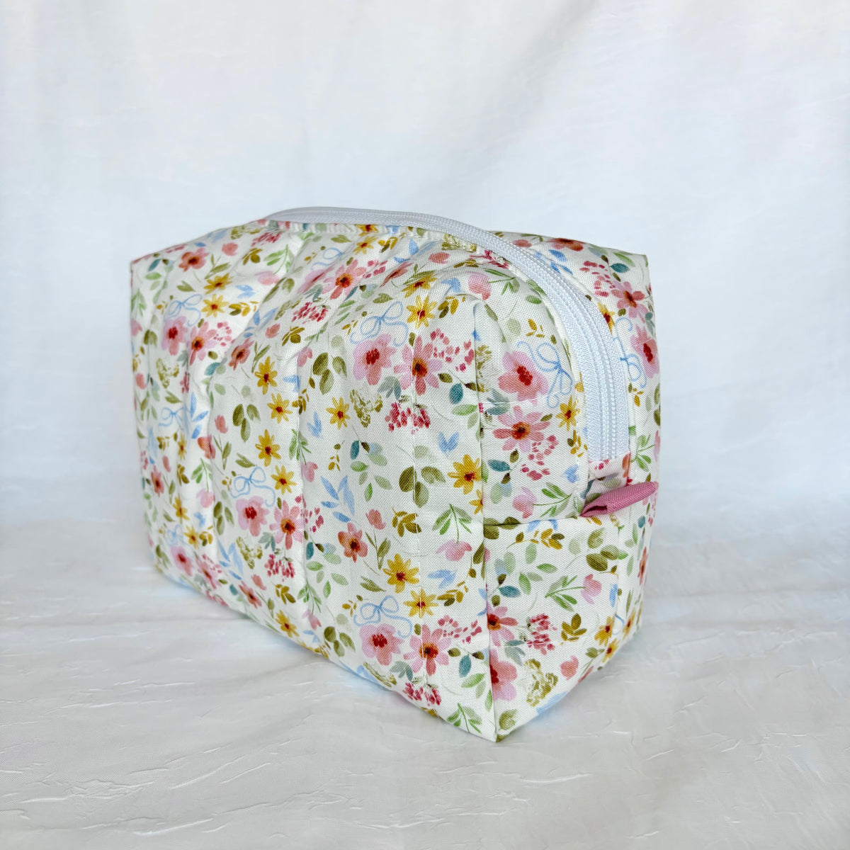 A rectangular zippered cosmetic pouch with a colorful floral print on a white background, featuring pink, yellow, and blue flowers with green leaves, and a white zipper.