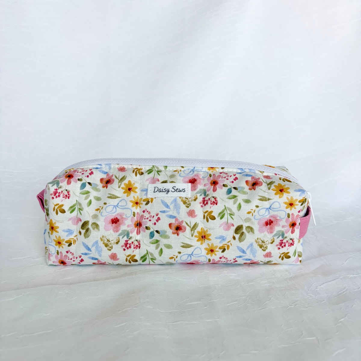 A fabric zippered pouch with a colorful floral print on a white background, featuring a small white label that reads 'Daily Sews'.