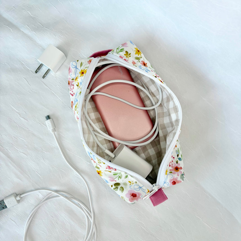 A floral and gingham travel pouch shown from above on a white textured fabric surface, with a pink rectangular item inside and white charging cables and a white wall charger nearby.
