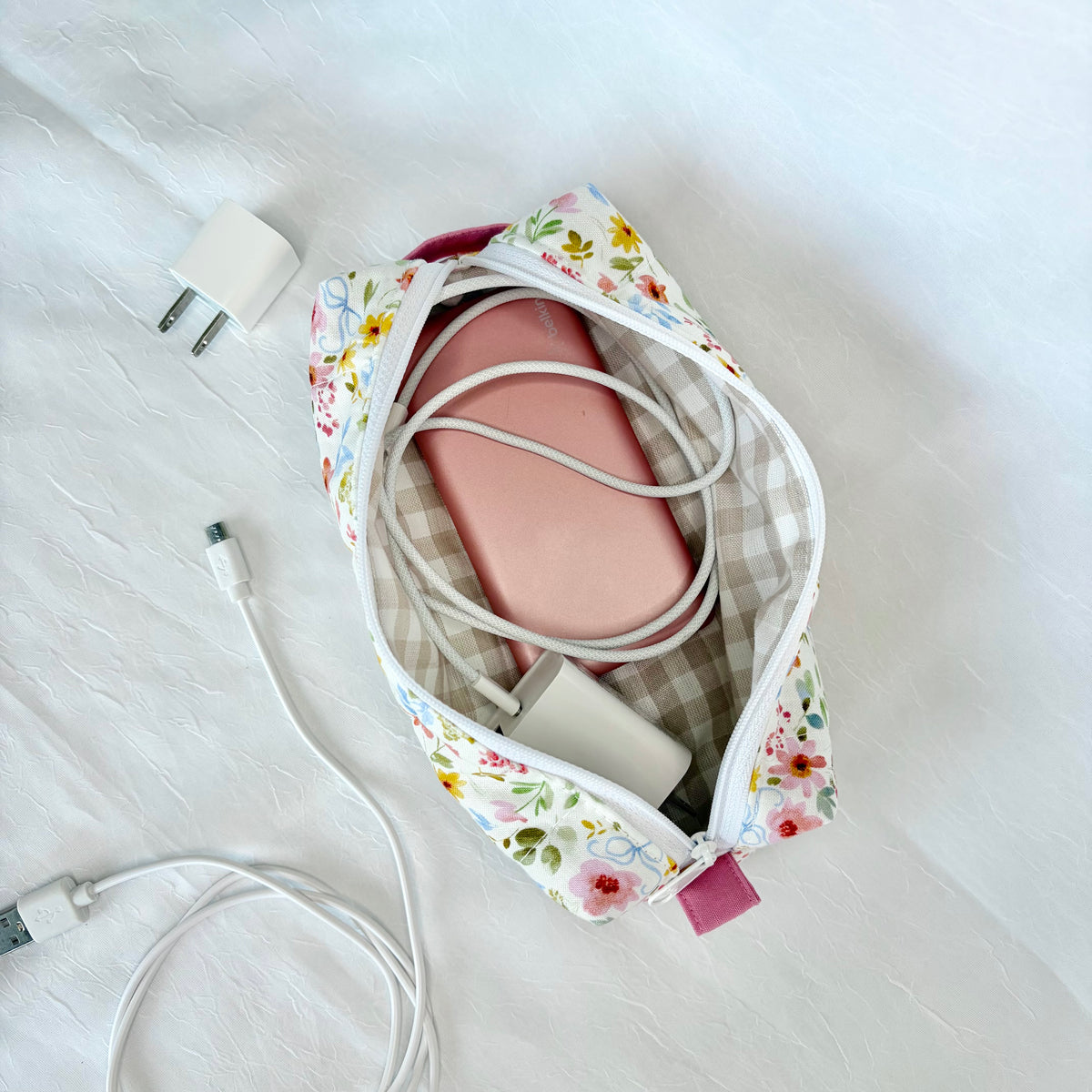 A floral and gingham travel pouch shown from above on a white textured fabric surface, with a pink rectangular item inside and white charging cables and a white wall charger nearby.