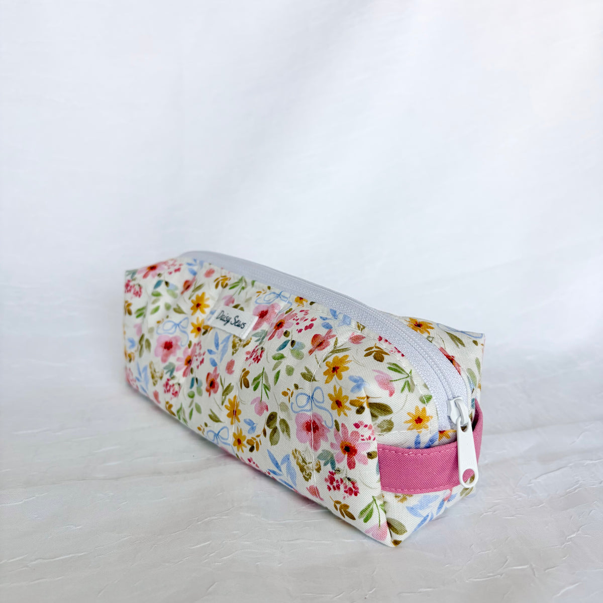 A fabric pencil case with a colorful floral print on a white background, featuring a silver zipper and a pink fabric loop handle.