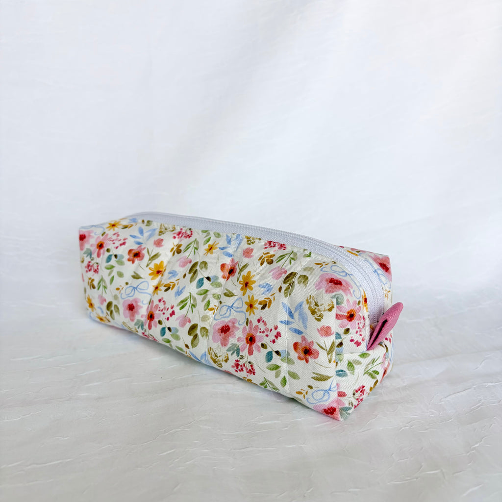 A rectangular zippered pencil case with a white background and a colorful floral print, featuring a pink pull tab on the zipper.