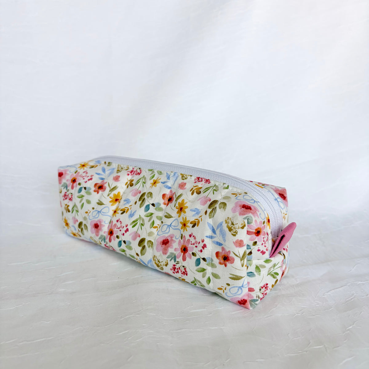 A rectangular zippered pencil case with a white background and a colorful floral print, featuring a pink pull tab on the zipper.