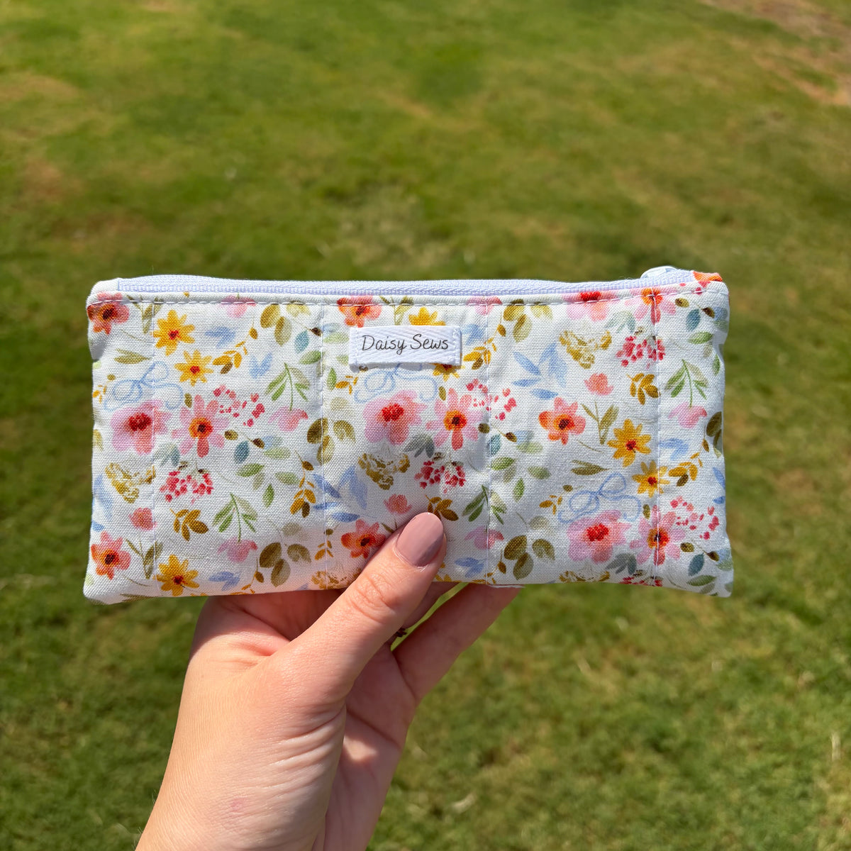 A hand holding a small zippered pouch with a white background and a colorful floral print, featuring a 'Daisy Sews' label, set against a blurred green grass background.