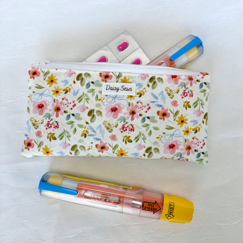 A floral zippered pouch with a white label reading 'Daisy Sews' lies on a white textured surface, partially filled with an epipen and pink pills. A pink and blue pen is placed in front of the pouch.