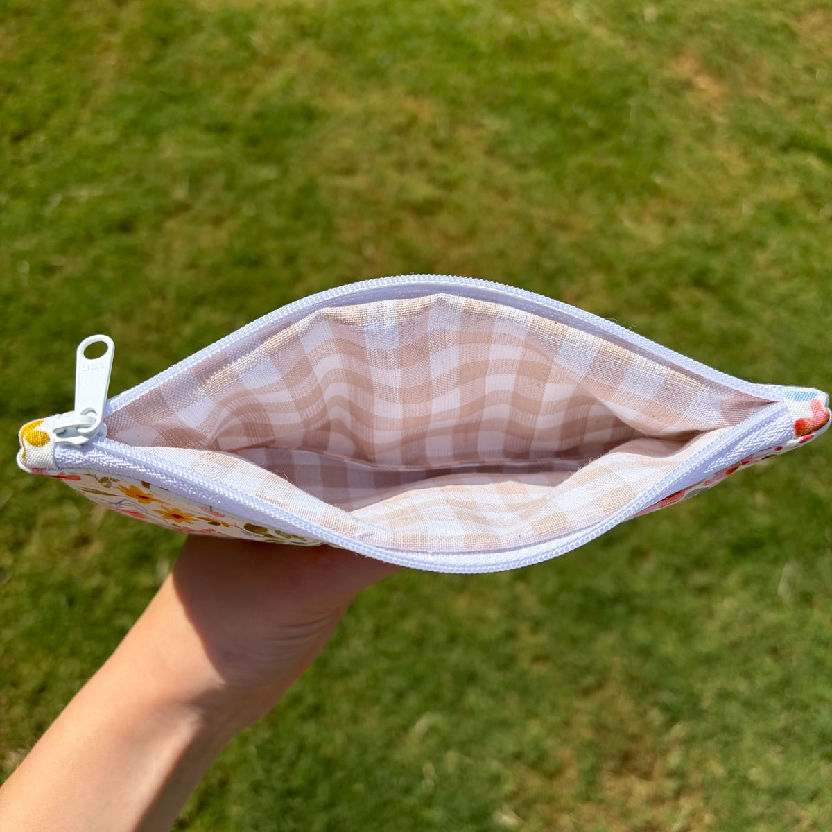 A hand holding open a small zip pouch outdoors on a grassy lawn. The pouch has a white and pale pink gingham lining and a white zipper pull.