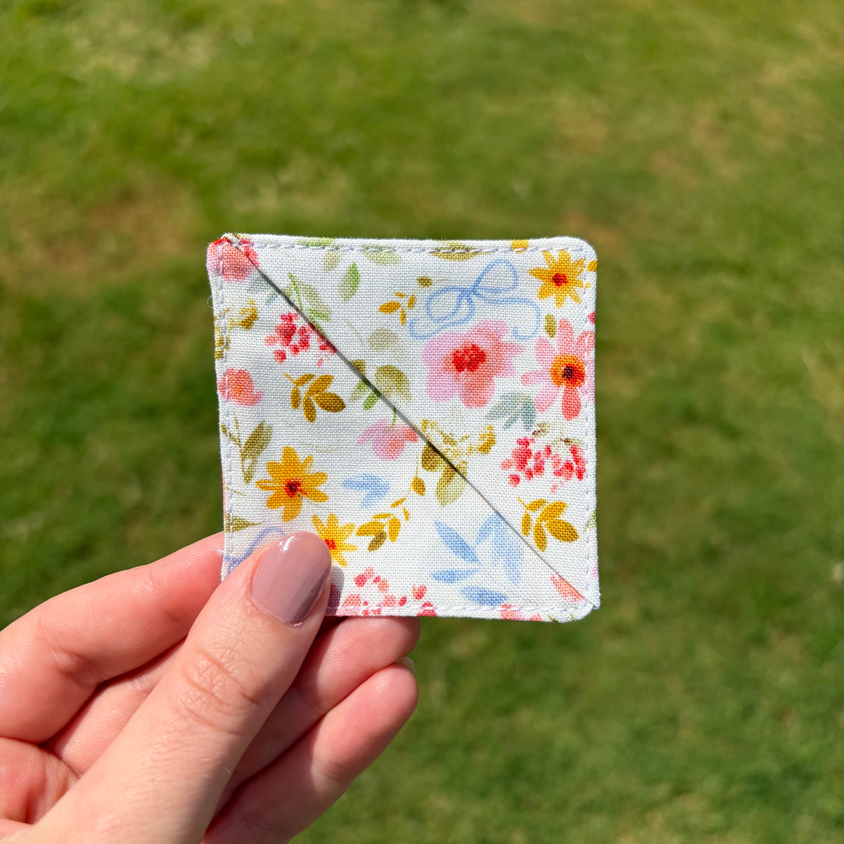 A hand holding a small square fabric patch with a white background and a colorful floral print, photographed outdoors against a blurred green grass background.