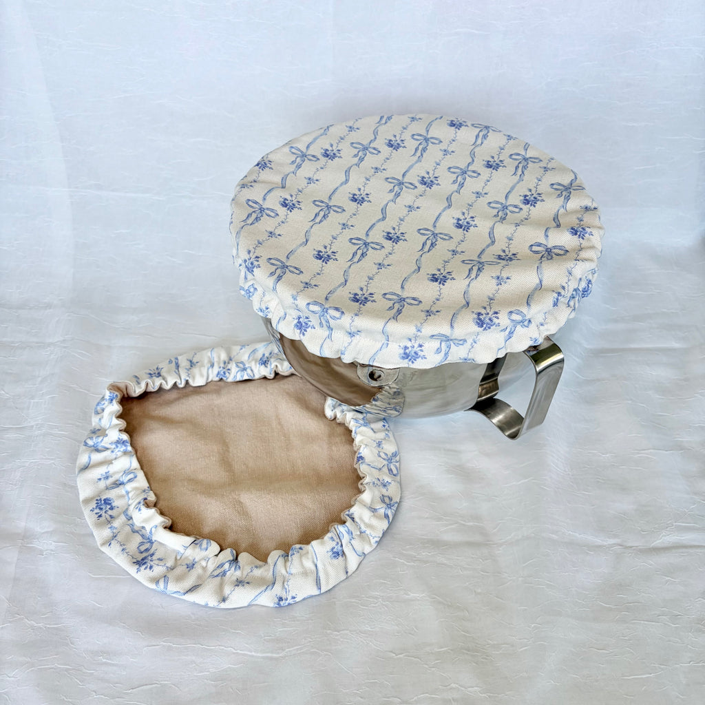 white cover with blue bows design on silver bowl with white background. tan fabric next to it