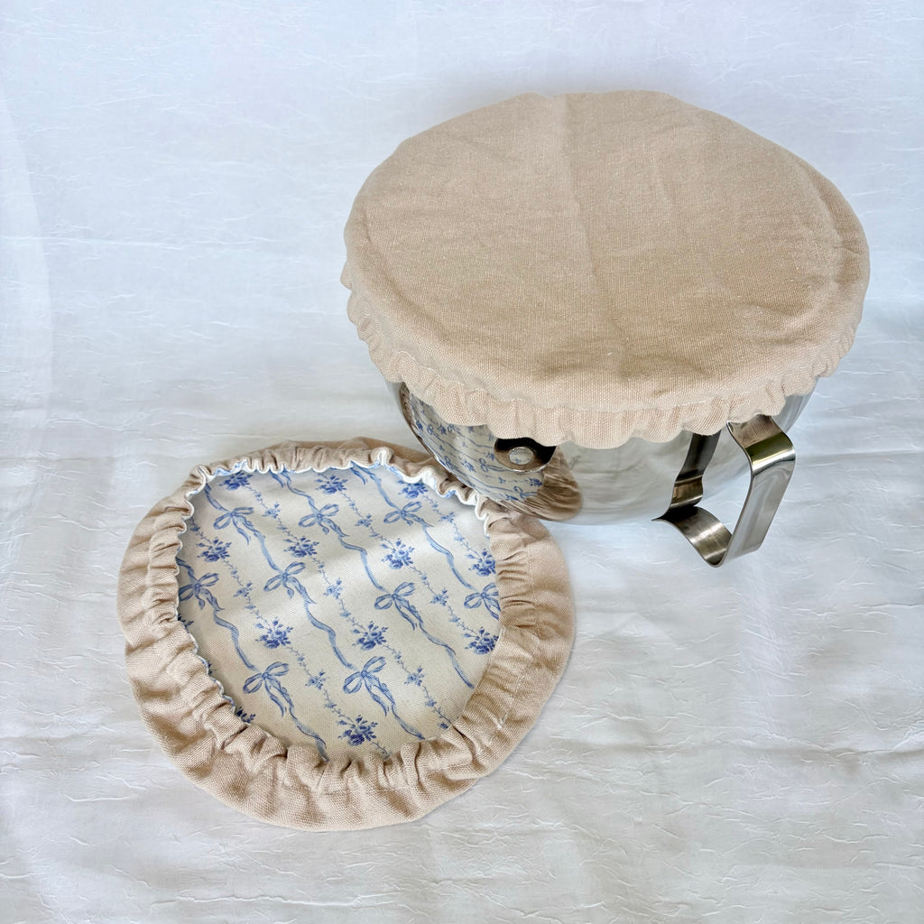 tan cover on silve bowl with white background. Blue bow fabric next to it