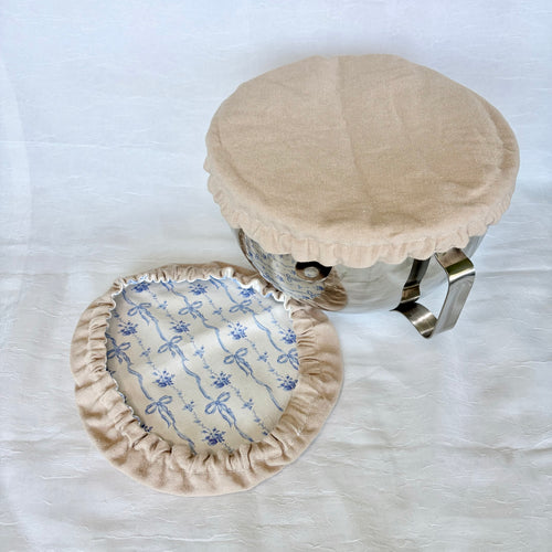 tan cover on silve bowl with white background. Blue bow fabric next to it