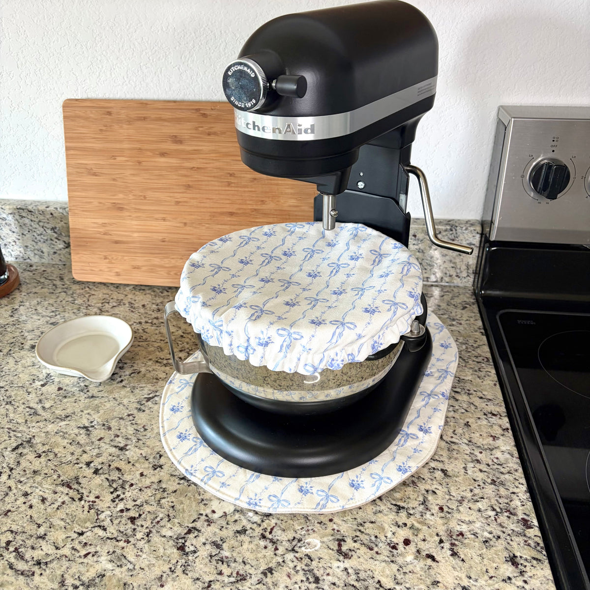 A black KitchenAid stand mixer with a white and blue floral bow-patterned dust cover on the bowl, set on a matching mat on a granite kitchen countertop next to a wooden cutting board and stove.