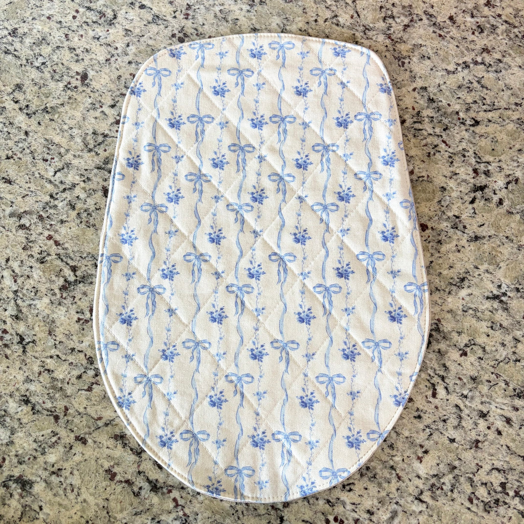 An oval quilted mat with a blue and white floral bow pattern, laid flat on a speckled granite countertop. head tilt size