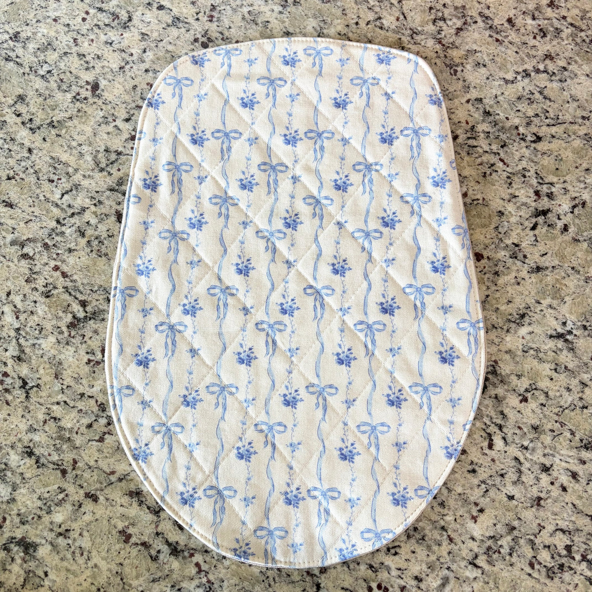 An oval quilted mat with a blue and white floral bow pattern, laid flat on a speckled granite countertop. head tilt size