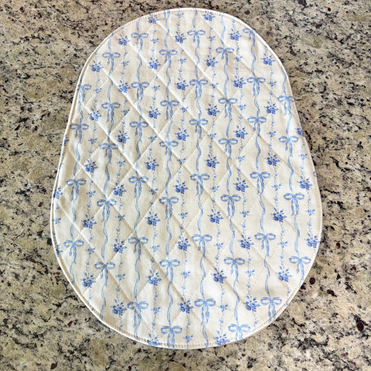 An oval quilted mat with a blue and white floral bow pattern, laid flat on a speckled granite countertop. bowl lift size