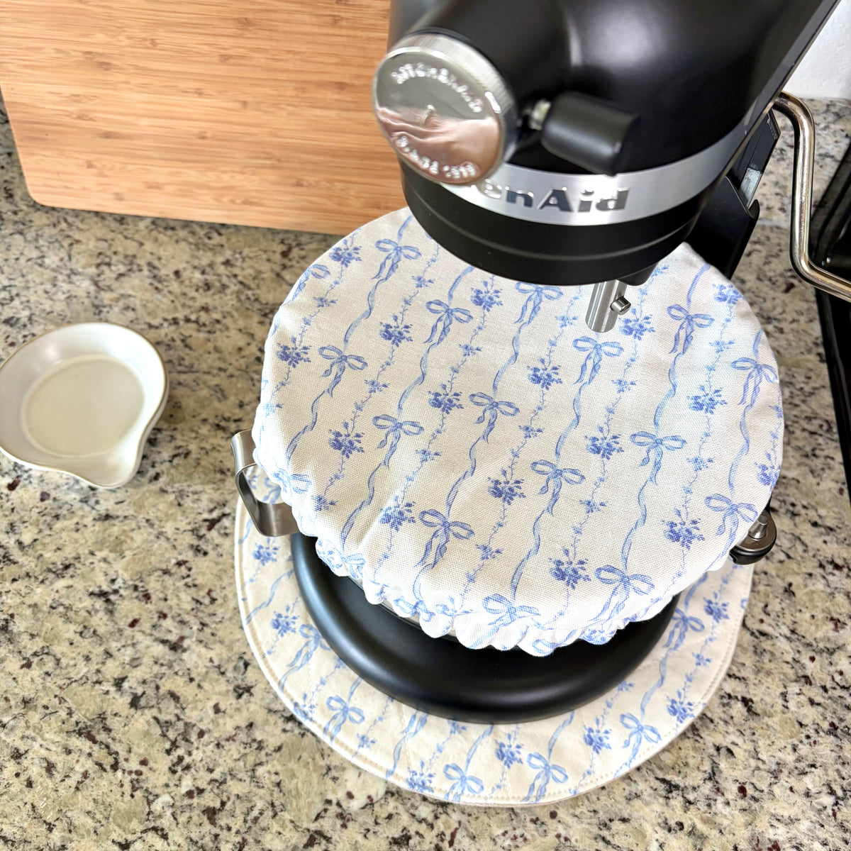 A close-up of a black KitchenAid stand mixer with a white fabric cover featuring a blue bow and floral pattern, set on a matching mat on a granite countertop.