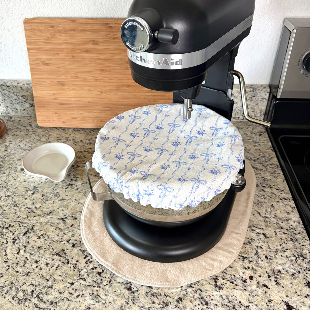 A black KitchenAid stand mixer with a white fabric cover featuring a blue bow pattern, sitting on a tan mat on a granite kitchen countertop.