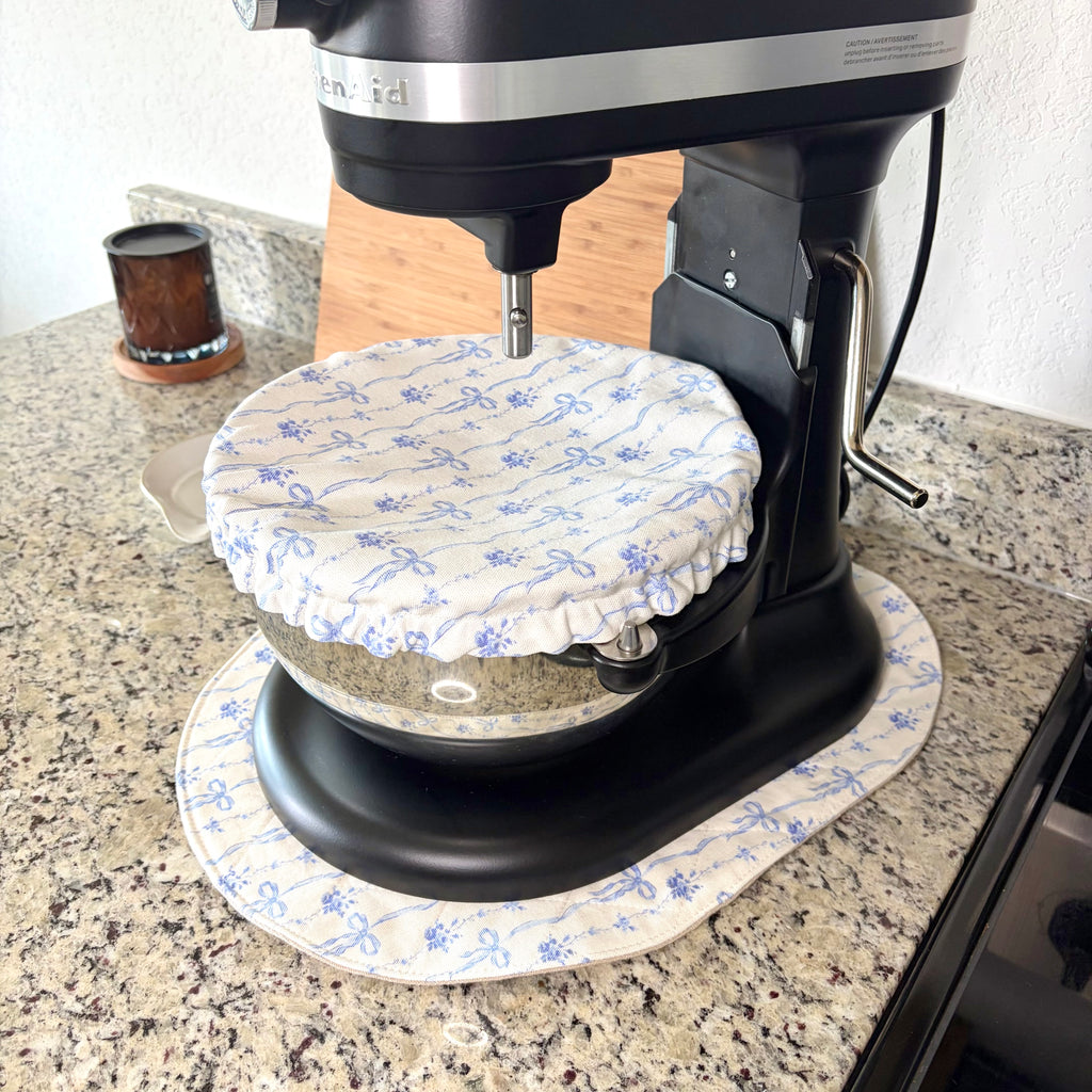 A black stand mixer with a glass bowl covered by a blue and white patterned fabric dust cover, sitting on a granite kitchen countertop.