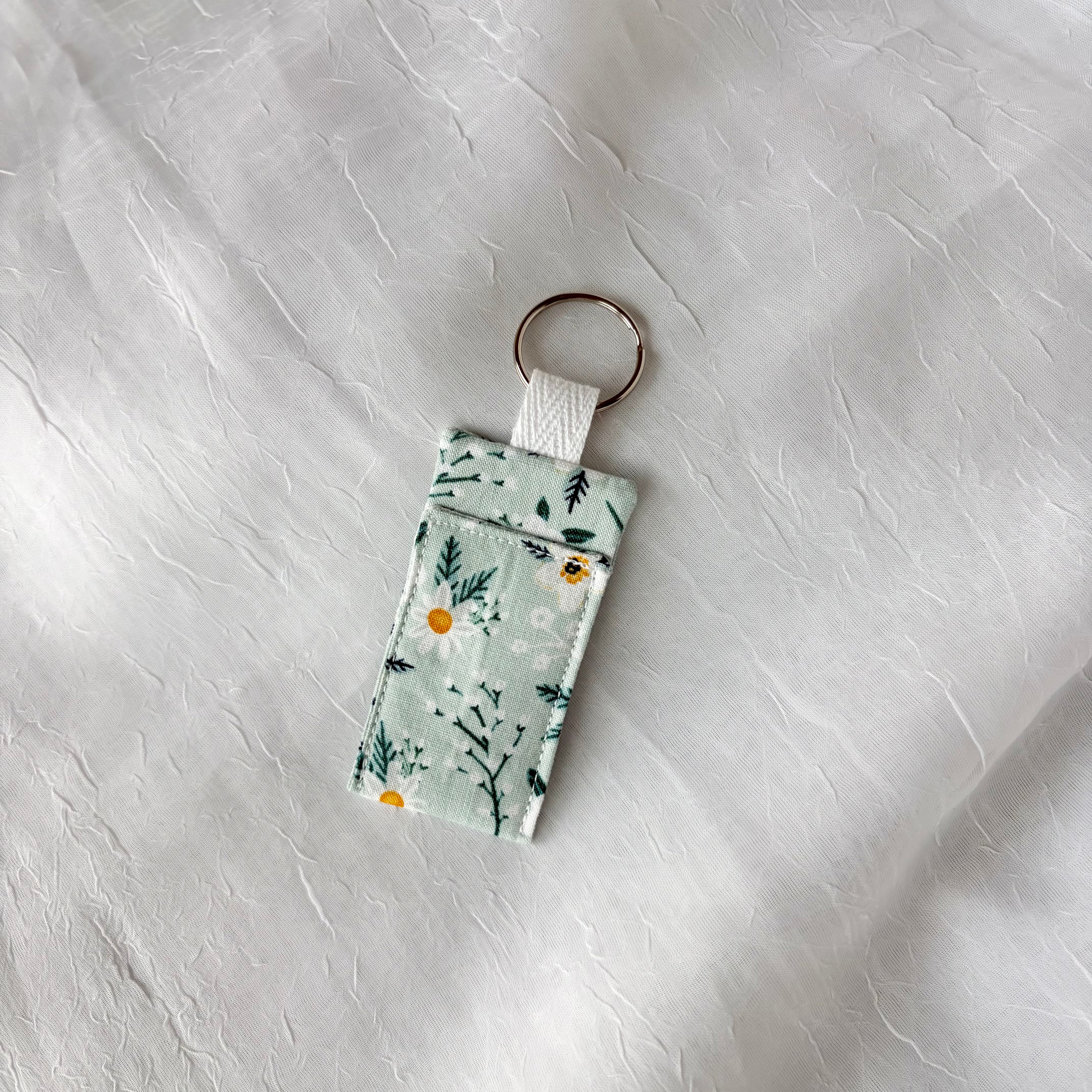 White flowers with yellow centers and dark teal accents on light blue fabric colored chapstick holder with keychain ring on a textured gray background