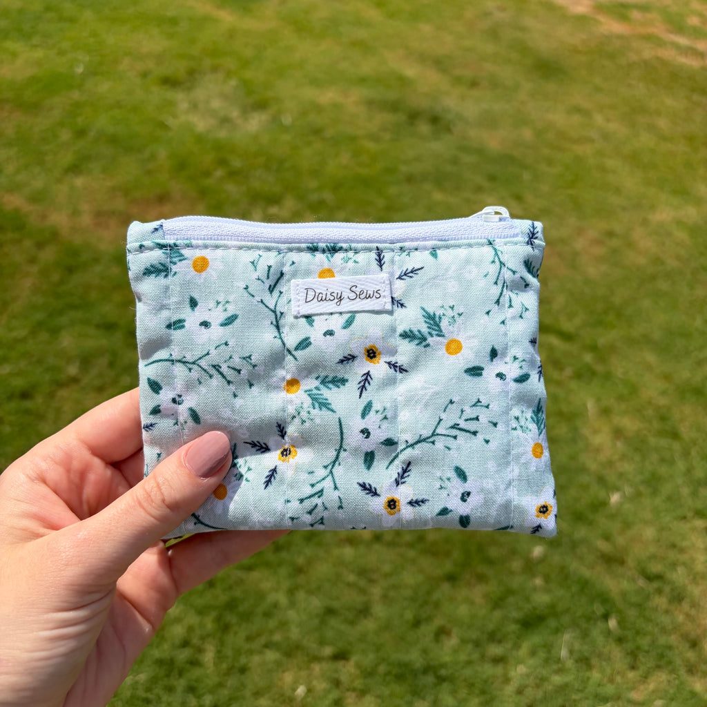 A hand holding a small zippered pouch with a light blue floral print, featuring a white label that reads 'Daisy Sews', set against a blurred green grass background.