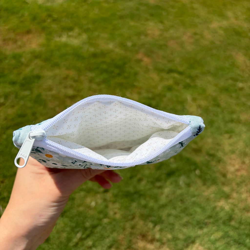 A hand holding an open zippered pouch with a white interior and a light blue floral exterior, set against a blurred green grass background.