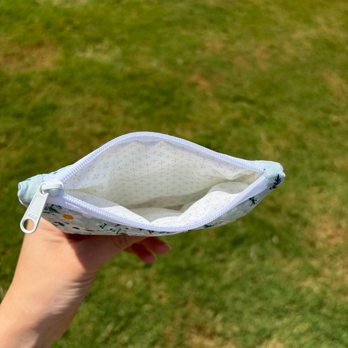 A hand holding an open zippered pouch with a white interior and a light blue floral exterior, set against a blurred green grass background.