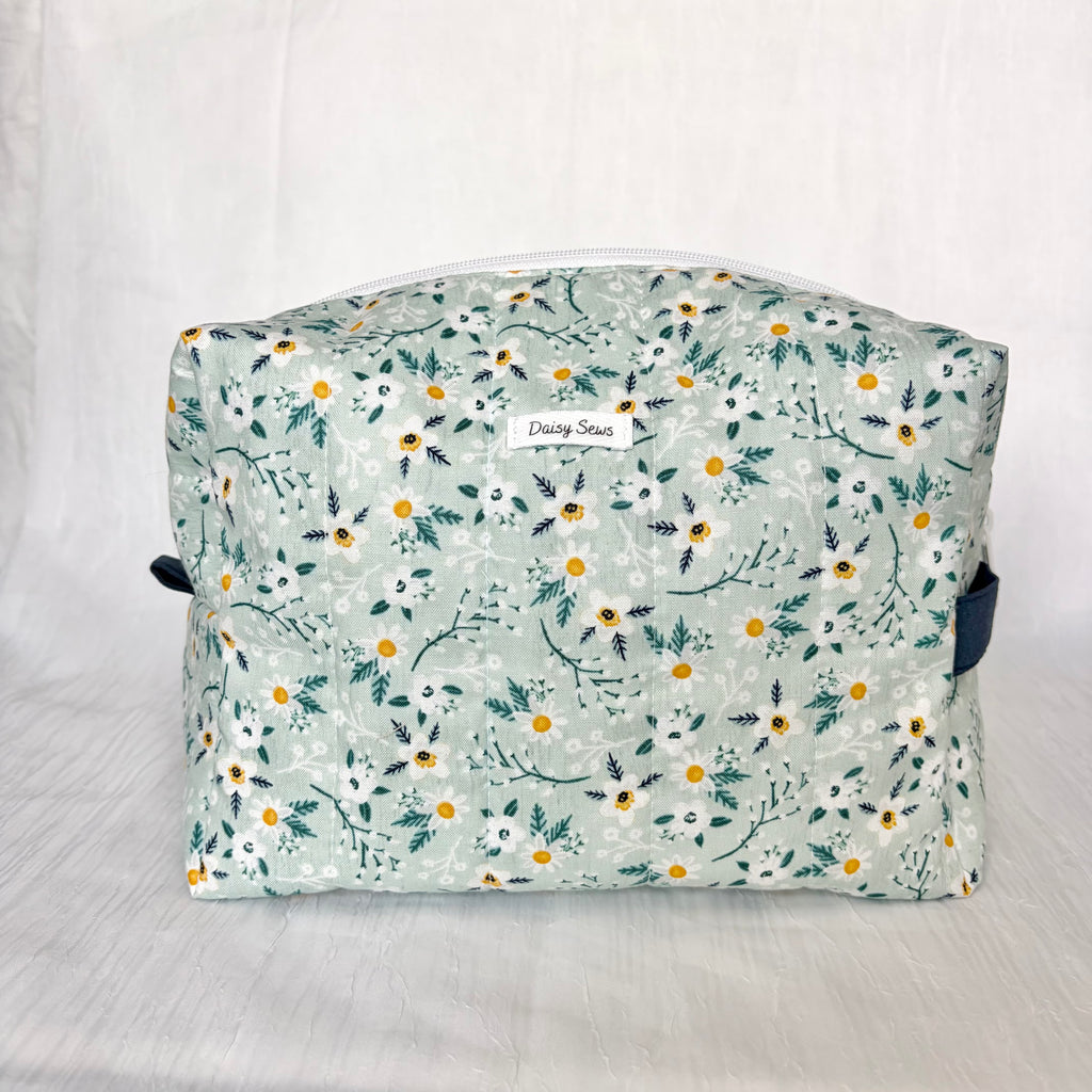 A quilted fabric cosmetic bag with a floral print in sage green, white, and yellow, featuring a white zipper and a small 'Daisy Sews' brand label on the front.