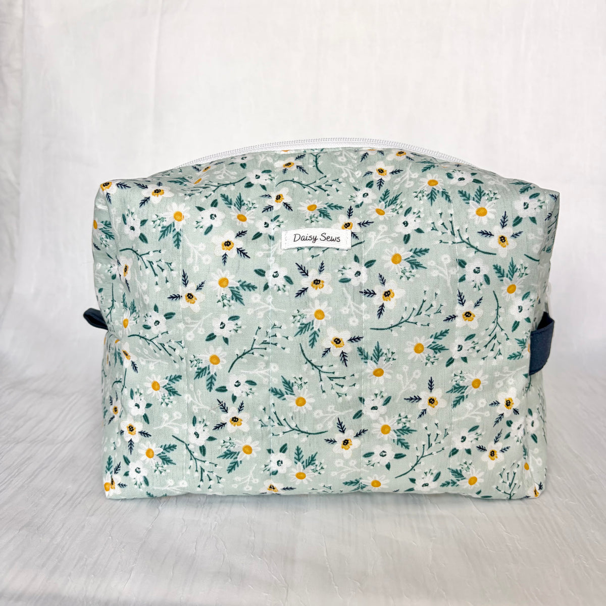 A quilted fabric cosmetic bag with a floral print in sage green, white, and yellow, featuring a white zipper and a small 'Daisy Sews' brand label on the front.